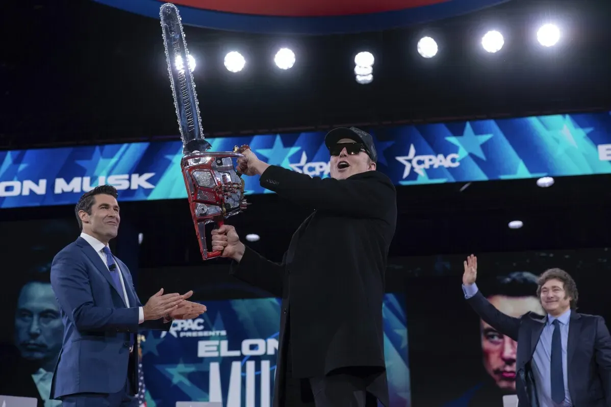 إيلون ماسك يحمل منشارًا مزخرفًا خلال مؤتمر العمل السياسي المحافظ، بينما يبتسم الحضور، مع خلفية تحمل شعار المؤتمر.