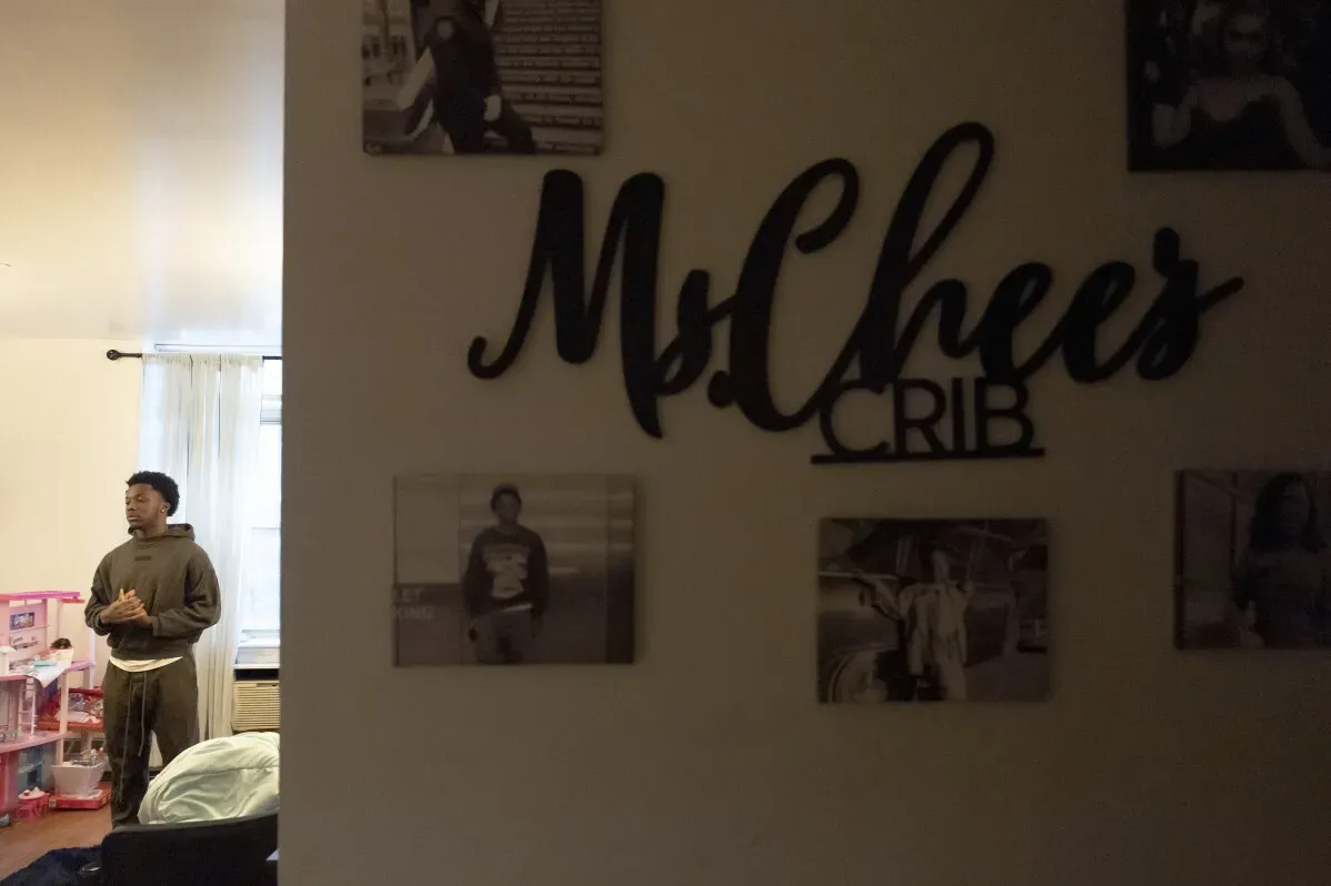 شاب يقف في غرفة مع ديكور منزلي، بينما يظهر على الحائط اسم "MsChees CRIB" مع صور عائلية. توضح الصورة الأثر الشخصي للأحداث المؤسفة.
