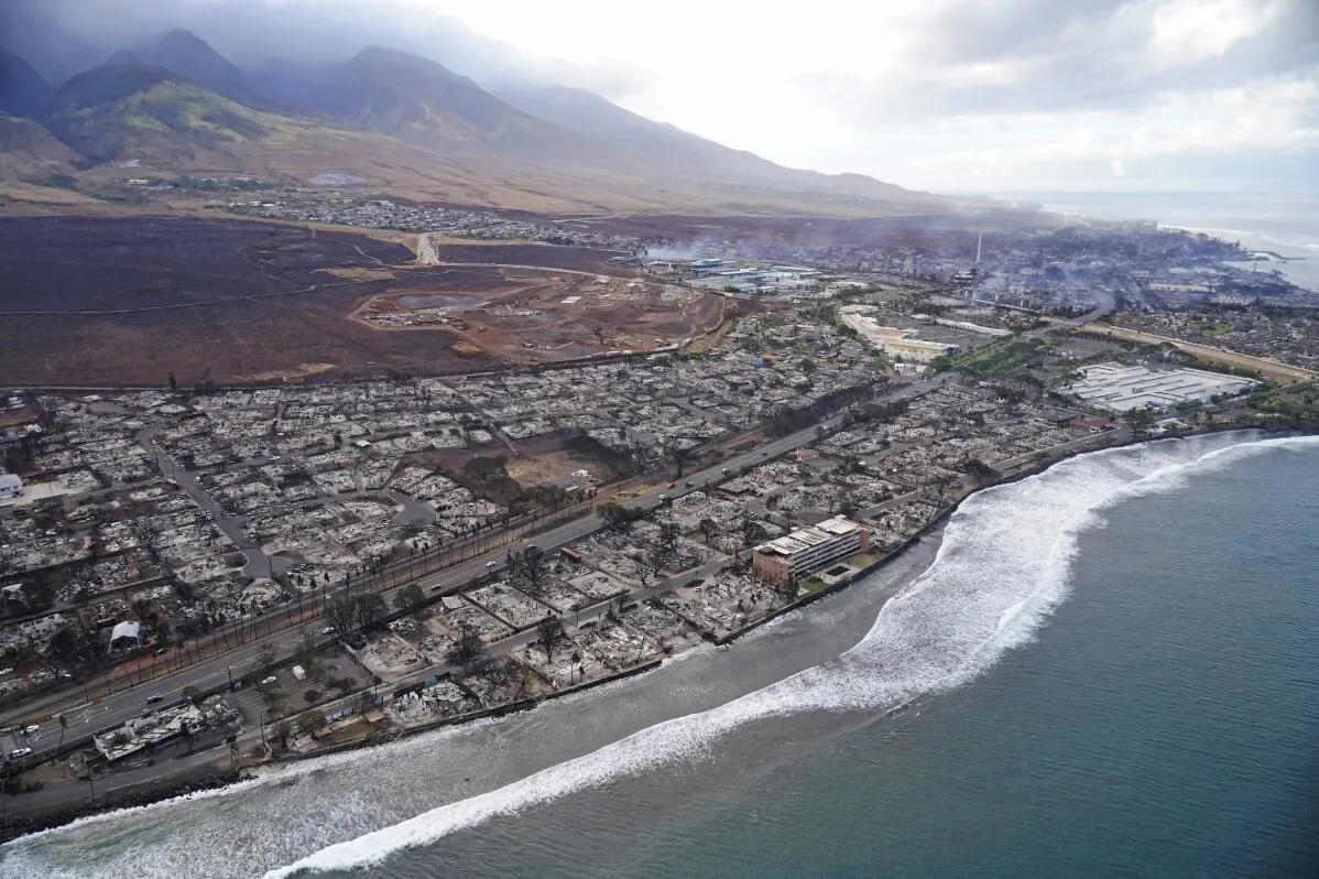 صورة جوية تُظهر الدمار الناتج عن حرائق الغابات في لاهاينا، هاواي، مع بيوت مدمرة وخط ساحلي متضرر، مما يعكس impacts جسيمة على الضحايا والمجتمع.