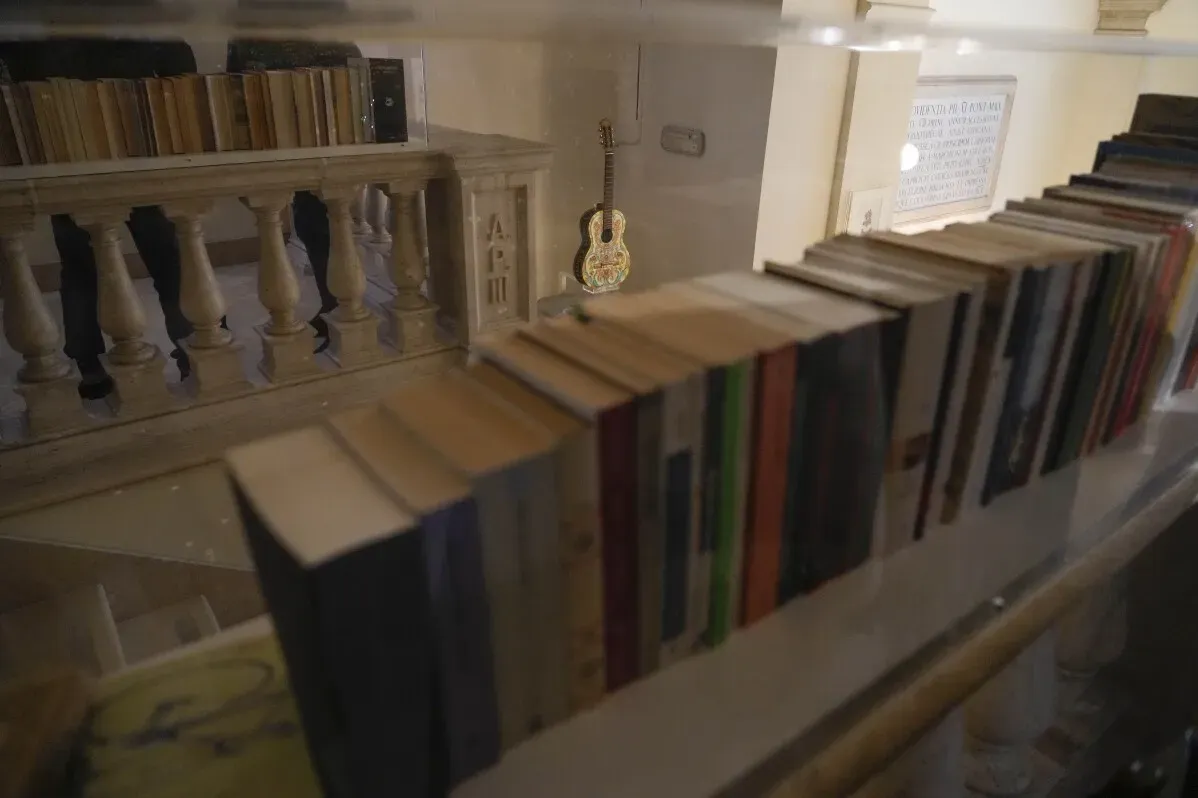 عرض لمكتبة الفاتيكان يتضمن مجموعة من الكتب المنسقة بشكل متقن، مع ظهور آلة موسيقية في الخلفية، تعكس التراث والفن المعاصر في المعرض.