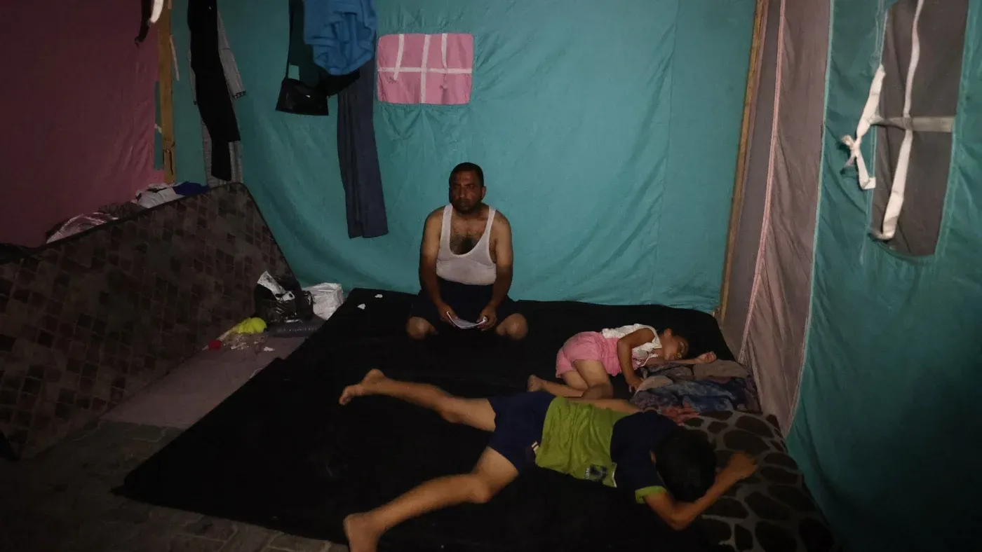رجل يجلس في خيمة بسيطة مع طفلين نائمين على الأرض، تعكس ظروف المعيشة الصعبة في غزة خلال الحرب والتهجير.