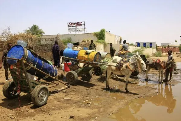 تظهر الصورة مجموعة من الأشخاص في منطقة أم درمان بالسودان، يجرون عربات محملة براميل مياه، وسط ظروف صعبة.