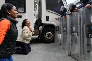 امرأة ترتدي سترة سوداء تجثو أمام رجال الشرطة الذين يرتدون دروعًا، بينما تعبر عن مشاعر الخوف والقلق في سياق الأوضاع السياسية المتوترة في فنزويلا.