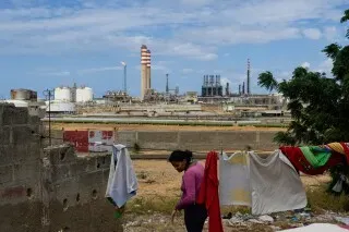 مشهد من كاراكاس، فنزويلا، يظهر امرأة تسير بجوار مصنع للنفط، مع أعمدة الدخان في الخلفية، مما يعكس الوضع الاقتصادي والسياسي المتقلب في البلاد.