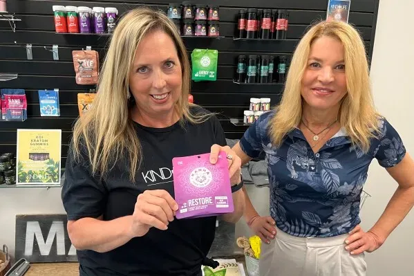 امرأتان في متجر Kind Life Dispensary، تحمل إحداهما علبة من علكات القنب، مع خلفية تحتوي على منتجات متنوعة.