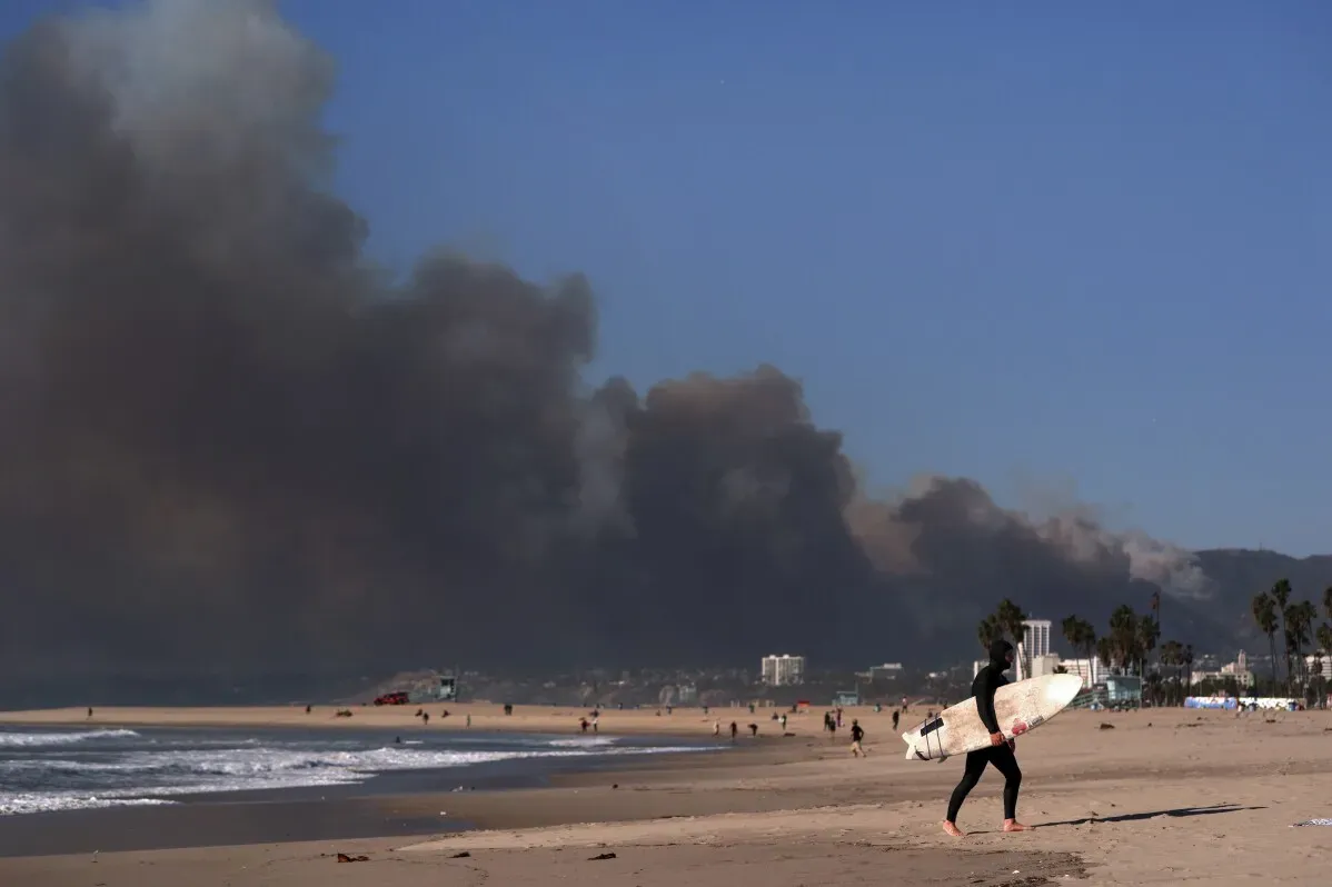 ساحل فينيسيا يظهر فيه شخص يحمل لوح تزلج، بينما تتصاعد سحب الدخان الكثيف من حرائق الغابات في الخلفية، مما يعكس تأثير الكوارث الطبيعية.