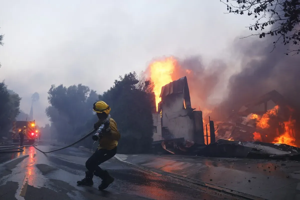 رجال الإطفاء يكافحون حريقًا هائلًا في باسيفيك باليساديس، مع ألسنة اللهب تتصاعد من المنازل المدمرة، وسط أجواء عاصفة.