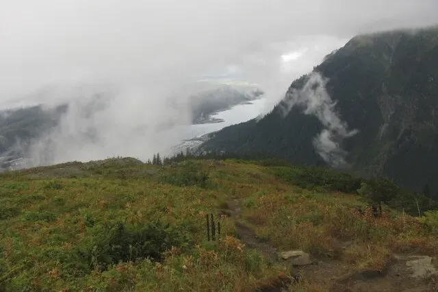 منظر طبيعي لجبل روبرتس في جونيو، ألاسكا، يظهر الضباب الكثيف والجبال المحيطة، مع مسار مشي يتجه نحو الأسفل.