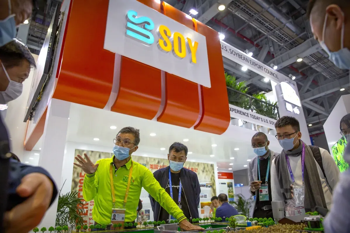 شخص يرتدي قميصًا أصفر يتحدث مع مجموعة من الأشخاص في معرض تجاري، مع وجود شعار "SOY" في الخلفية، مما يشير إلى موضوع التجارة الزراعية.