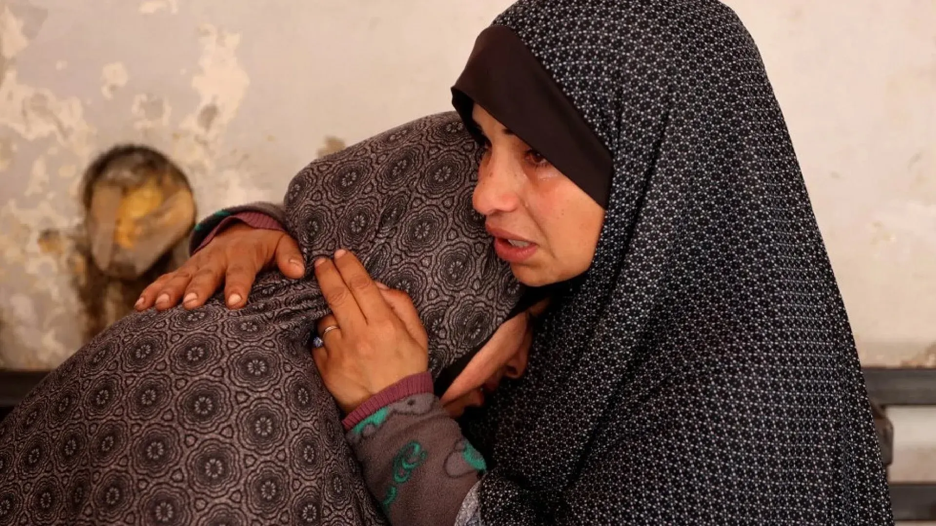 امرأتان ترتديان الحجاب، تتعانقان في لحظة حزن عميق، تعكسان تأثير الأزمات الإنسانية على النساء في غزة.