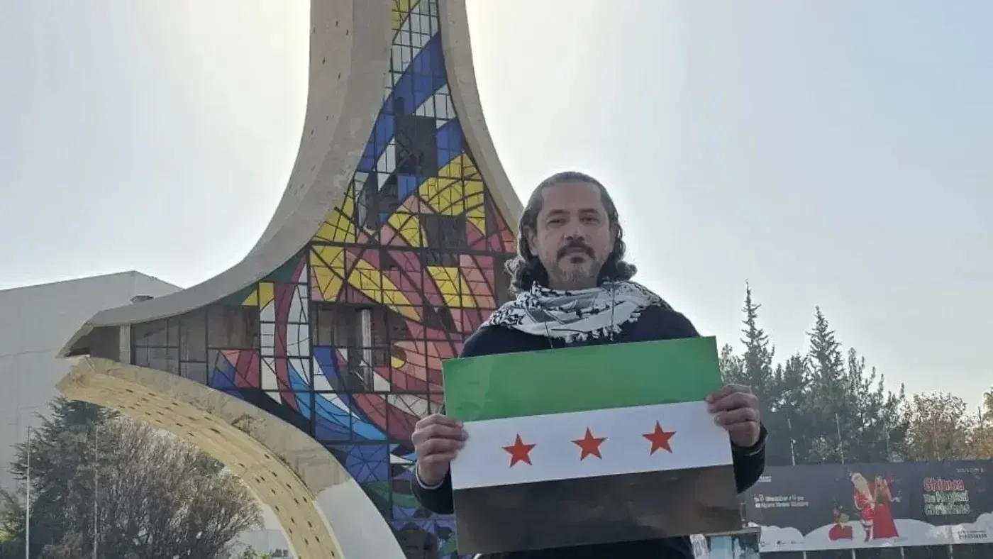 رجل يحمل علم الثورة السورية أمام مبنى مميز في لبنان، معبرًا عن دعمه للمعارضة السياسية في المنطقة.