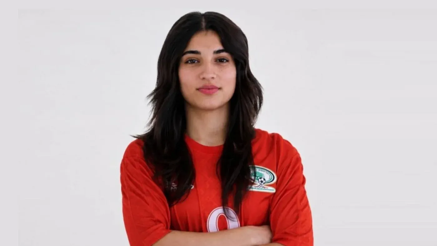 لاعبة كرة قدم فلسطينية ترتدي قميص المنتخب الوطني، تعبر عن القوة والعزيمة. الصورة تعكس معاناة الرياضيين الفلسطينيين في ظل الاعتقالات.