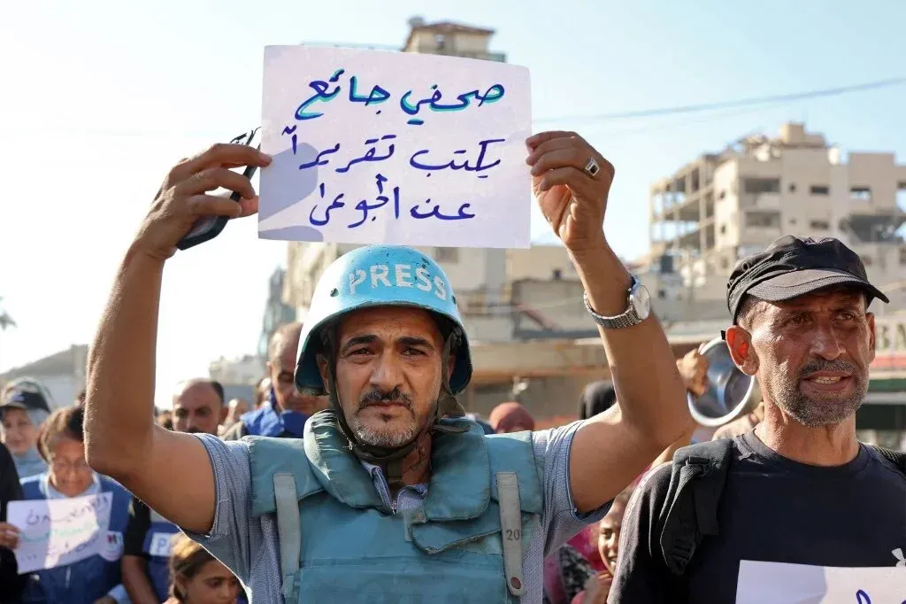 صحفي يرتدي خوذة ويحمل لافتة تطالب بالحرية للصحافة في غزة، مع وجود حشود من الناس في الخلفية.
