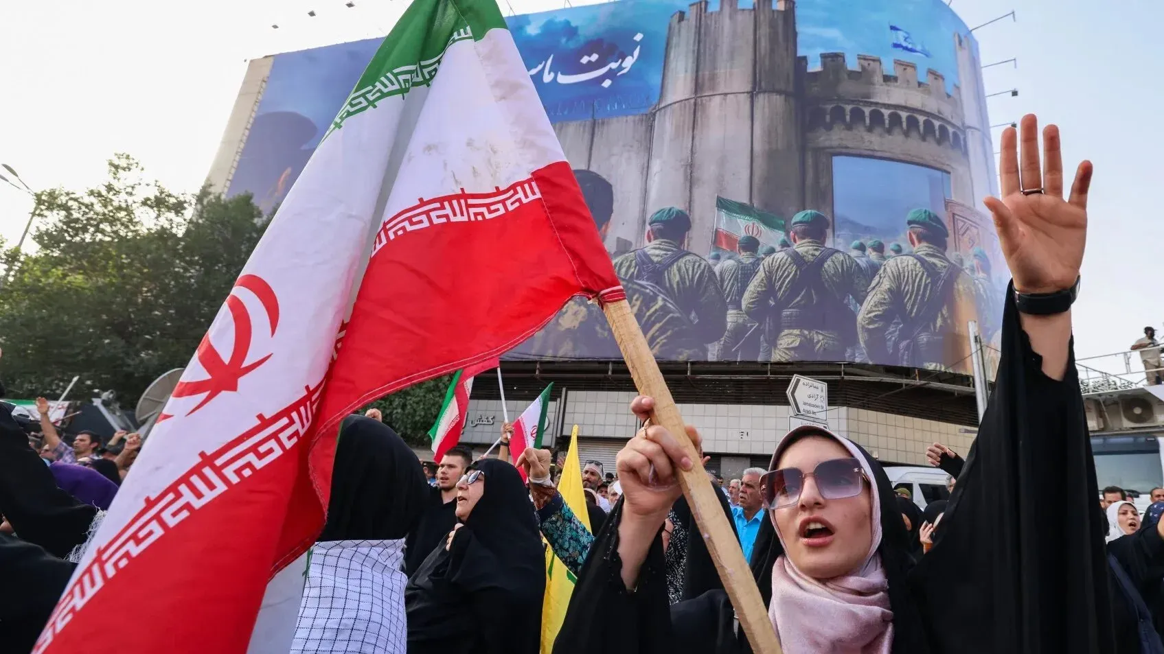 مظاهرة في إيران حيث تحمل امرأة علم البلاد وتظهر مشاعر قوية، مع خلفية لجدارية تمثل القوات الإيرانية.