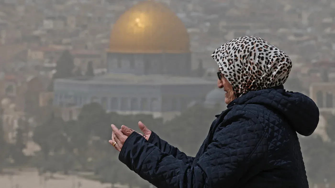 امرأة ترتدي حجابًا تقف وتدعو أمام قبة الصخرة في القدس، رمز المسجد الأقصى والتحديات التي تواجهه.