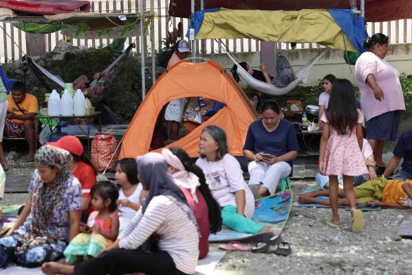 نازحون من زلزال الفلبين يجلسون في ملجأ مؤقت، مع خيمة برتقالية في الخلفية، يعكسون حالة الصدمة بعد الكارثة.