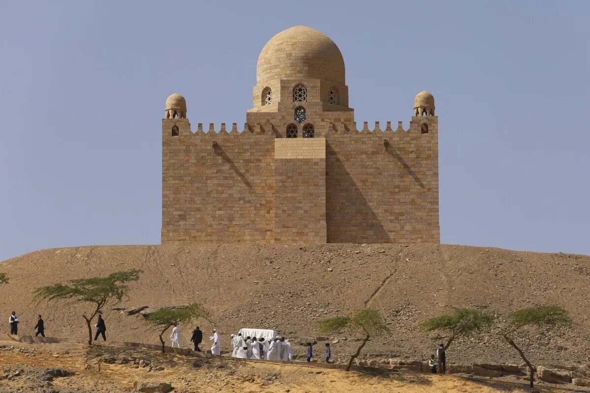 مراسم جنازة الآغا خان الرابع في أسوان، حيث يحمل المشيعون جثمانه الملفوف بكفن أبيض بالقرب من ضريح تاريخي.