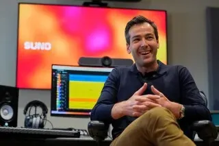 الرئيس التنفيذي لشركة Suno، ميكي شولمان، يجلس في مكتب استوديو التسجيل، محاطًا بأدوات الموسيقى والتكنولوجيا، أثناء تطوير أغنية جديدة باستخدام الذكاء الاصطناعي.