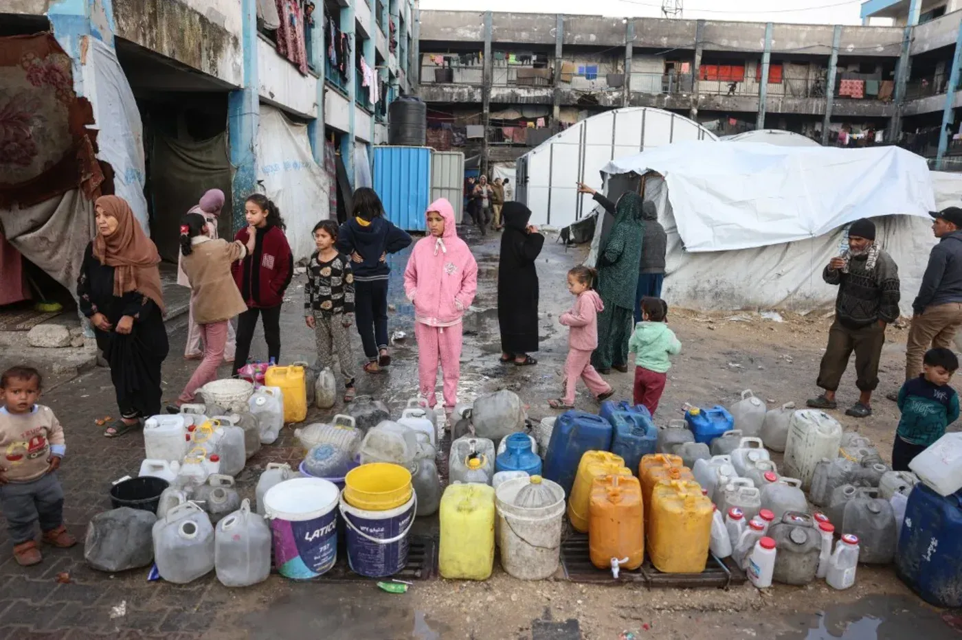 نساء وأطفال يقفون بجوار صف من حاويات المياه في مخيم للاجئين في غزة، مما يعكس الوضع الإنساني الصعب واحتياجات المساعدة الأساسية.
