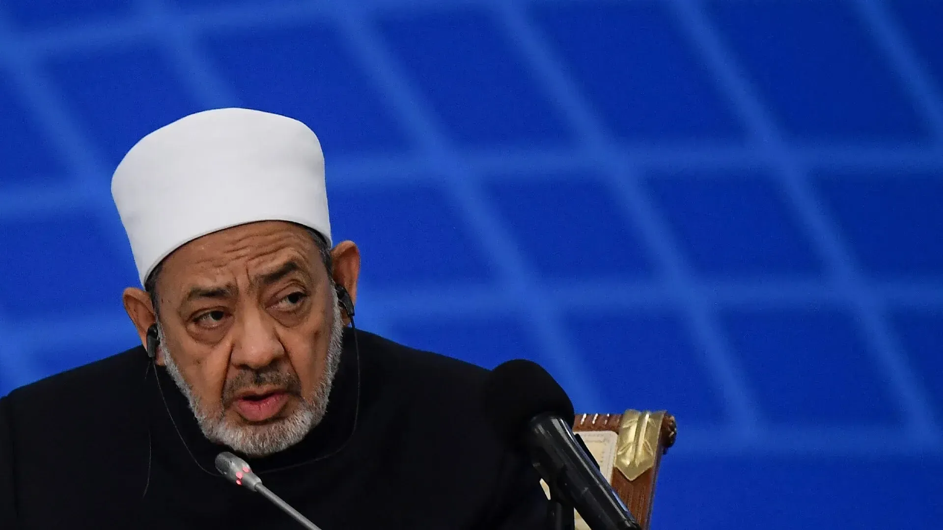 شيخ الأزهر أحمد الطيب يتحدث خلال مؤتمر، معبرًا عن قلقه بشأن الإبادة الجماعية في غزة، وسط خلفية زرقاء.