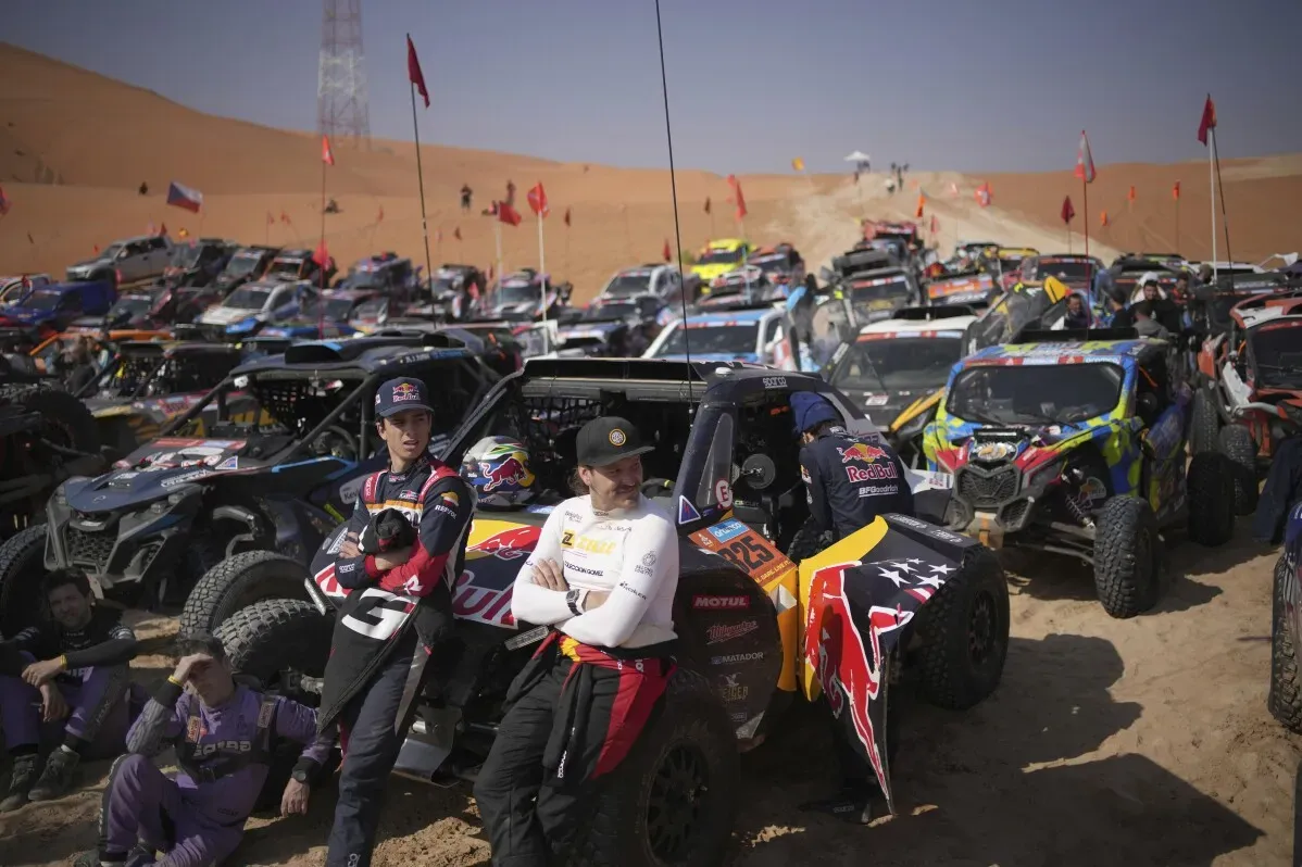 يظهر في الصورة مجموعة من سيارات الدفع الرباعي المشاركة في رالي داكار في الصحراء السعودية، مع سائقين يتحدثون في المقدمة.