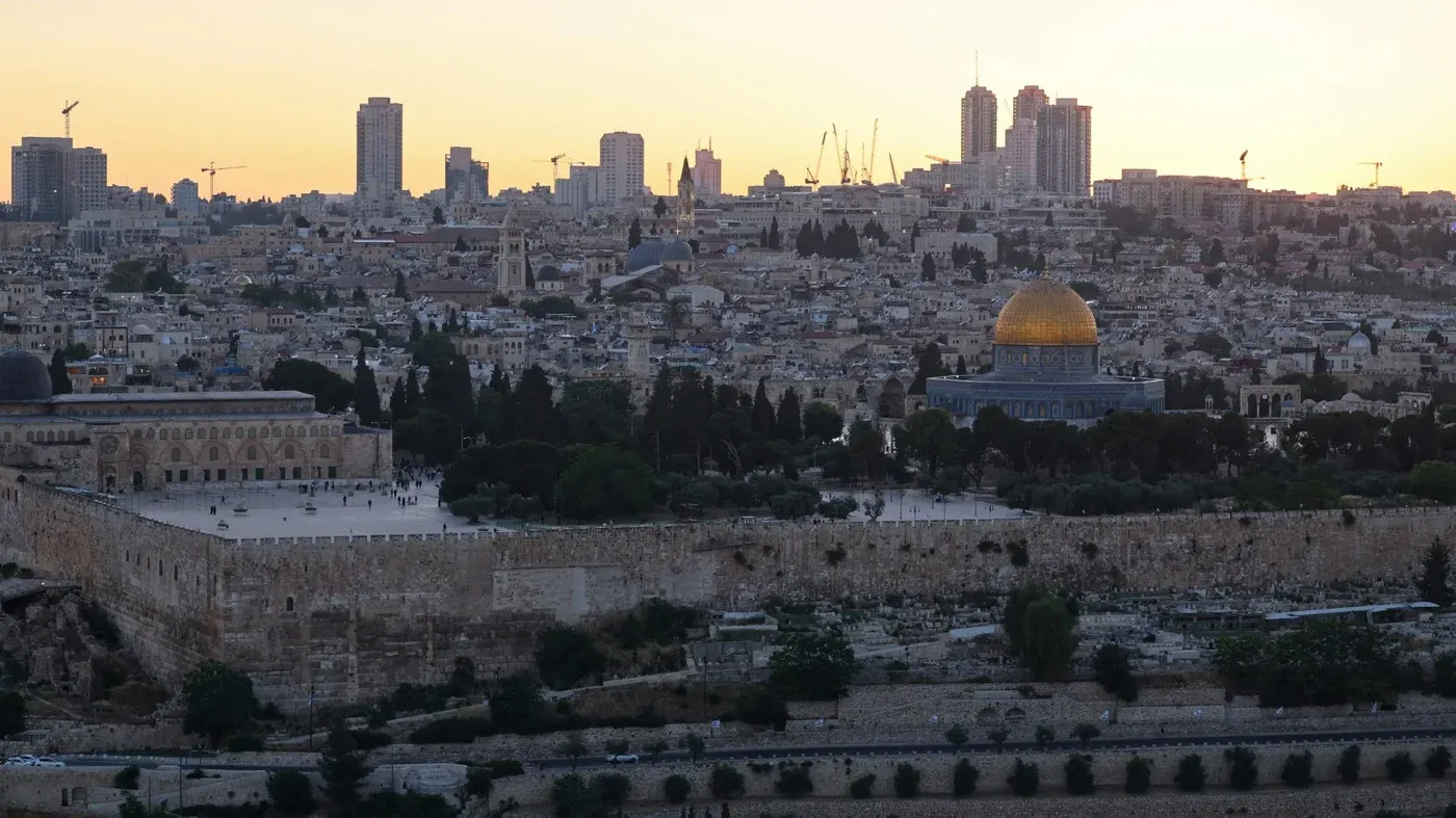 القدس القديمة تظهر في الغروب، مع قبة الصخرة البارزة في المقدمة، محاطة بمباني حديثة، تعكس الصراع بين التاريخ والحداثة.