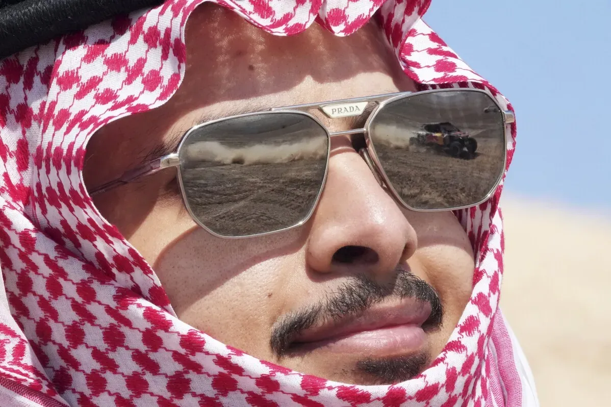 سائق يرتدي نظارات شمسية تعكس صورة مركبة في رالي داكار، يظهر في الخلفية صحراء المملكة العربية السعودية.