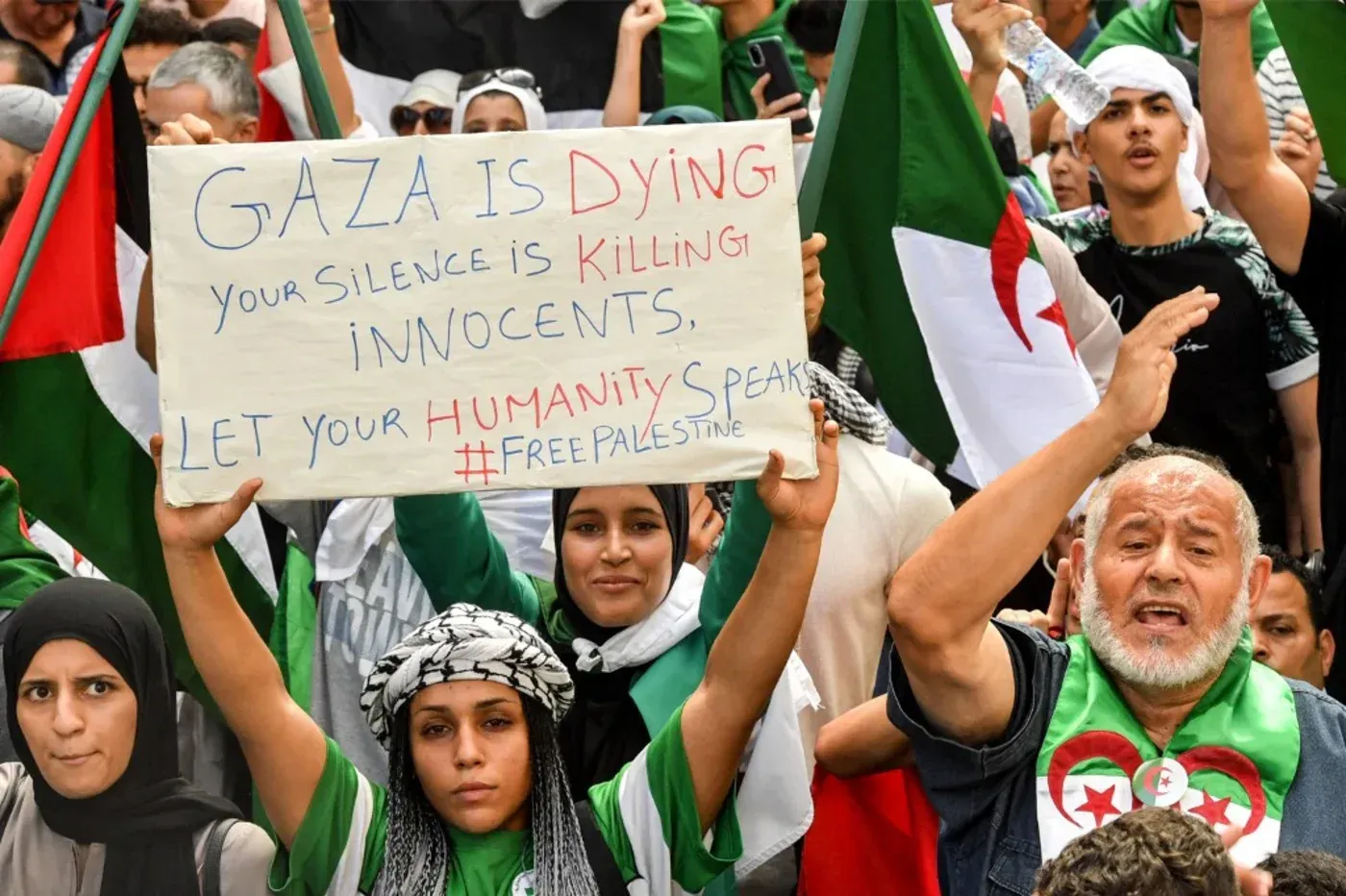 محتجون جزائريون يحملون لافتة تدعو لدعم غزة، معبرين عن تضامنهم مع الفلسطينيين وسط أعلام الجزائر، في سياق قمع المظاهرات.