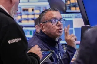 متداول في بورصة نيويورك يتحدث بحماس، محاط بشاشة تعرض بيانات السوق. تعكس تعابيره القلق بشأن تراجع الأسهم وأسعار السلع.