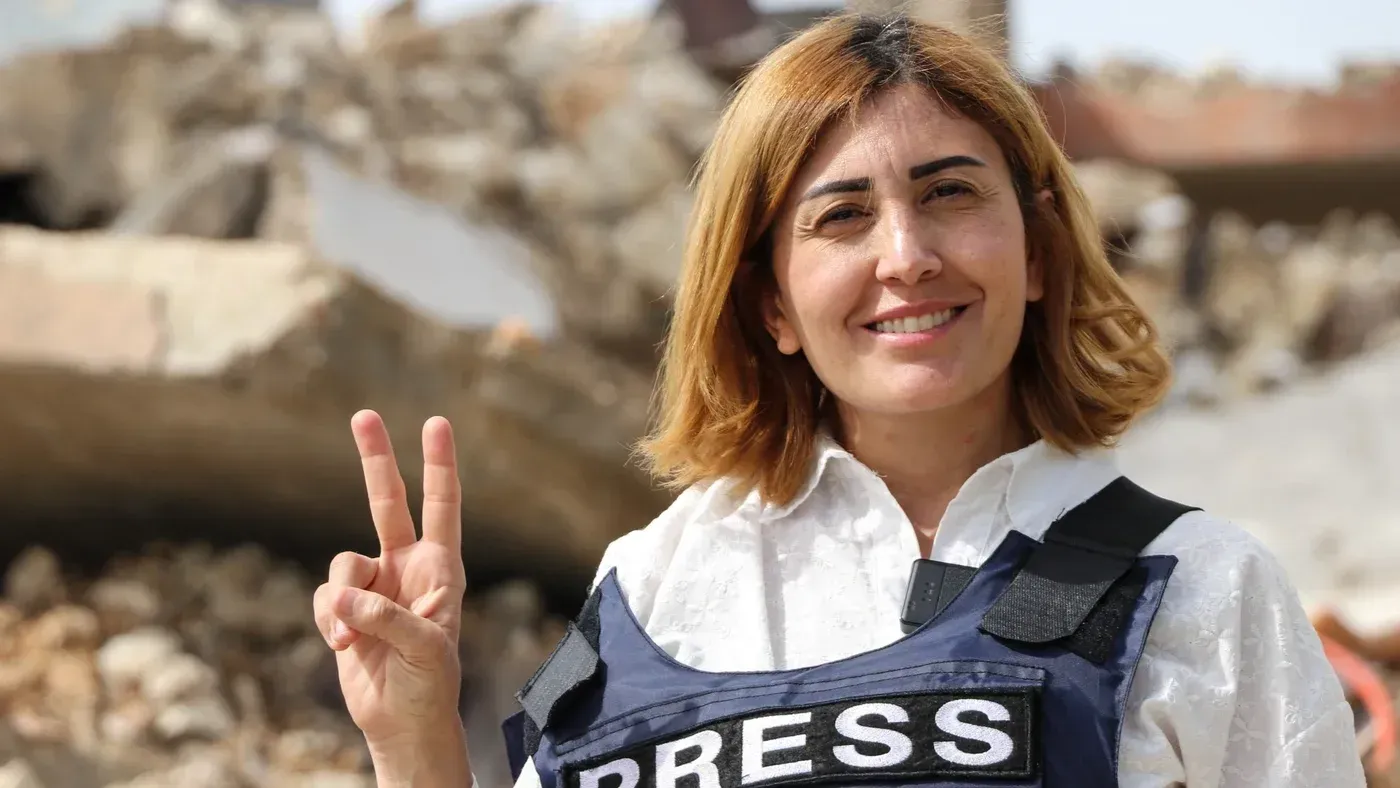 آمال خليل، الصحفية اللبنانية، مبتسمة وترتدي سترة تحمل علامة "صحافة"، ترفع إصبعها في إشارة النصر أمام أنقاض مبنى مدمر.
