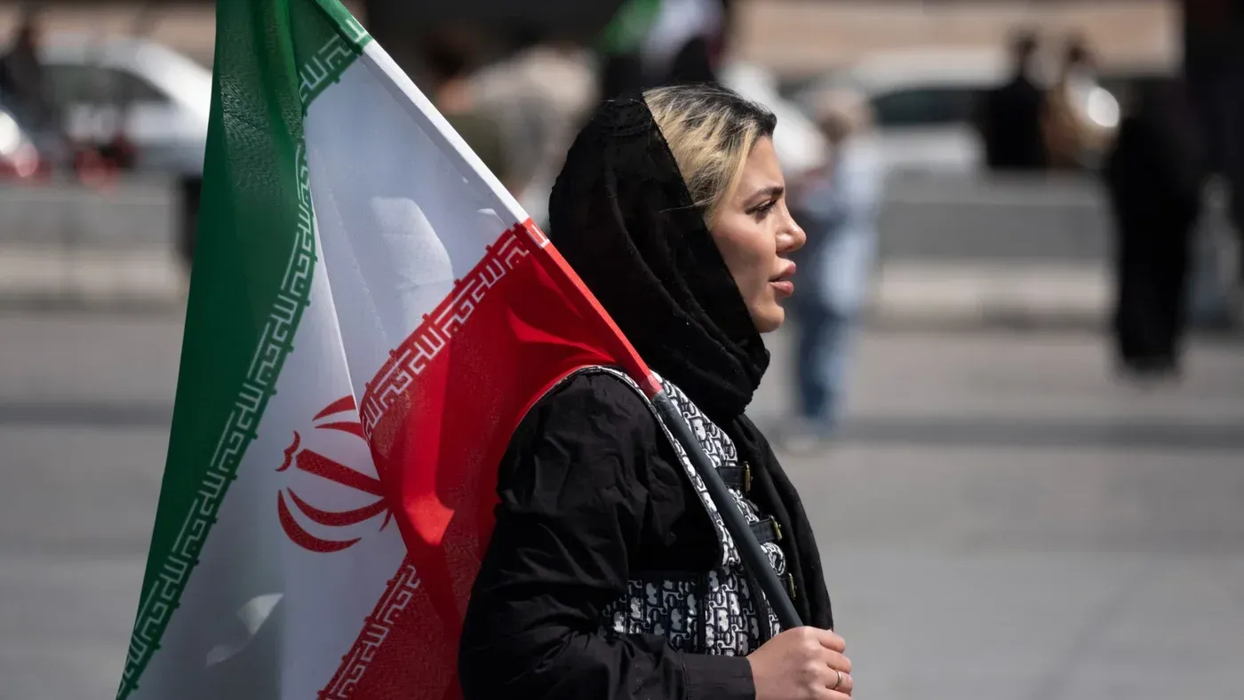 امرأة ترتدي حجابًا وتحمل علم إيران، تظهر في مشهد حضاري يعكس التغيرات الاجتماعية في طهران. تعكس الصورة تنوع الحياة اليومية في البلاد.
