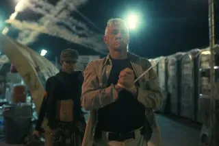 مشهد درامي من فيلم "المذنبون"، يظهر شخصية رئيسية تتقدم في بيئة عسكرية مضاءة بأضواء ساطعة، مع تأثيرات خلفية تعكس التوتر.