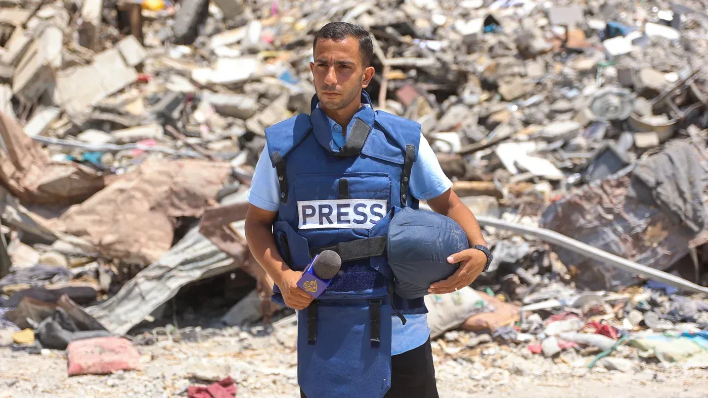 صحفي فلسطيني يرتدي سترة تحمل كلمة "صحافة" أمام أنقاض في غزة، يعكس التحديات التي يواجهها الإعلام في زمن الحرب.