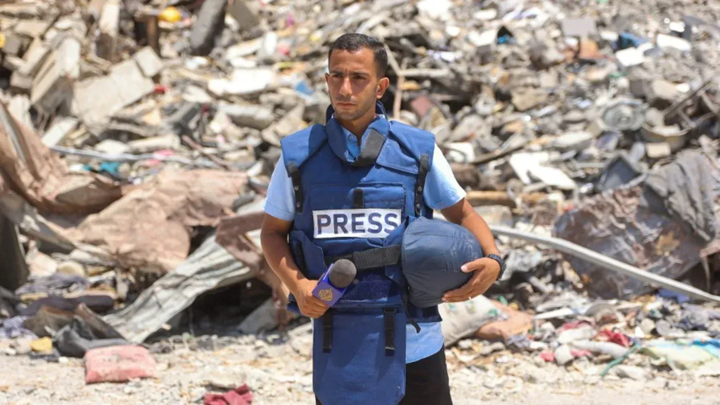 صحفي فلسطيني يرتدي سترة صحافة زرقاء، يقف وسط حطام في غزة، معبرًا عن التحدي والشجاعة في مواجهة الحرب.
