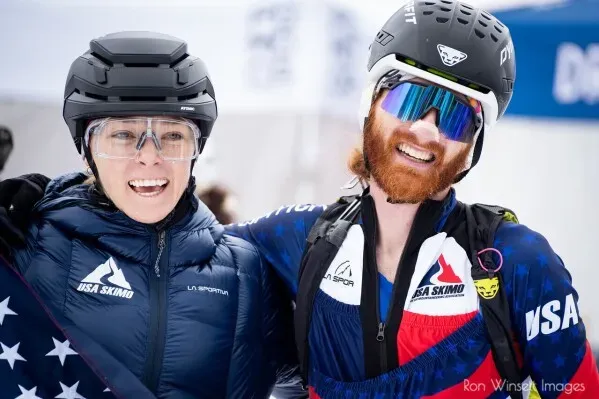 آنا جيبسون وكاميرون سميث مبتسمتان بعد سباق تزلج، يرتديان ملابس رياضية وخوذات، تعكسان روح التعاون في رياضة السكيمو.