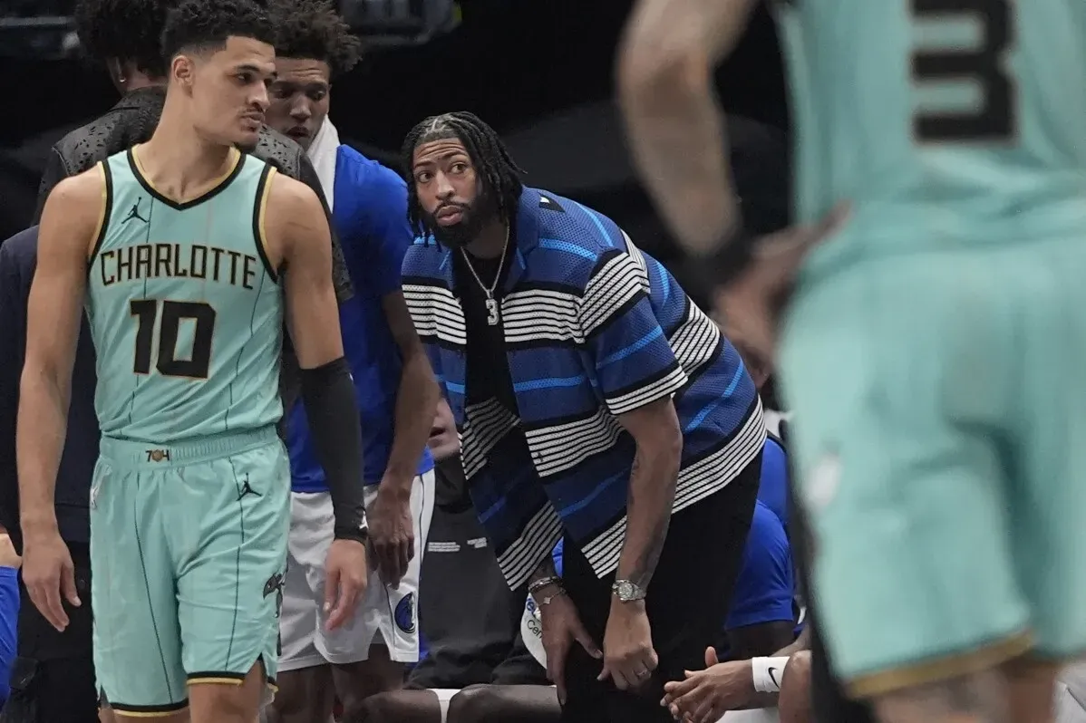 أنتوني ديفيس يتابع المباراة من مقاعد البدلاء، مرتديًا قميصًا مخططًا، بينما لاعب من فريق شارلوت يتجه نحو الملعب.