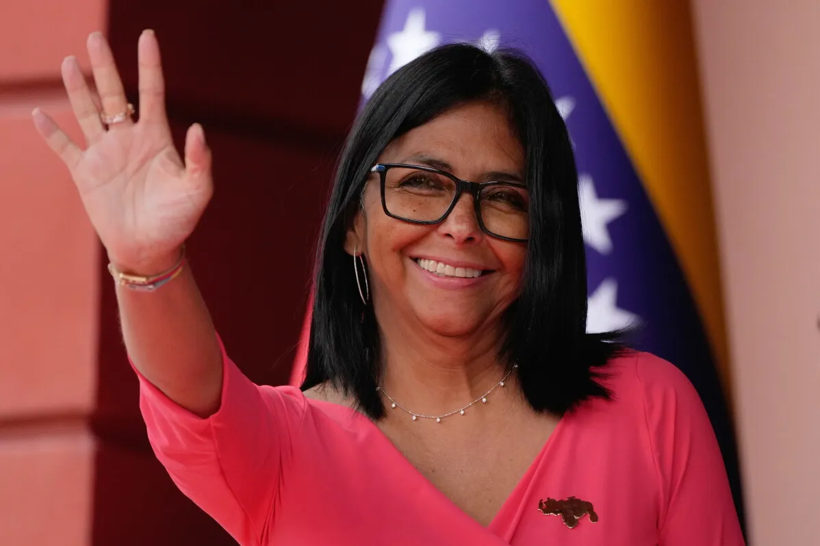 تظهر Delcy Rodríguez، الرئيسة الفنزويلية بالإنابة، مبتسمة وتلوح بيدها، مع العلم الفنزويلي خلفها، في سياق التوترات السياسية الحالية.