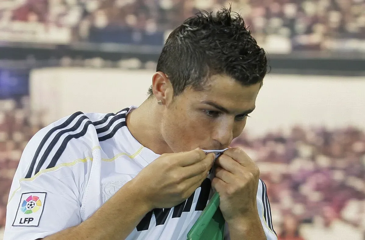 كريستيانو رونالدو، يرتدي قميص ريال مدريد، يقبّل الشعار في لحظة احتفالية، مع خلفية جماهيرية تعكس شغف كرة القدم.