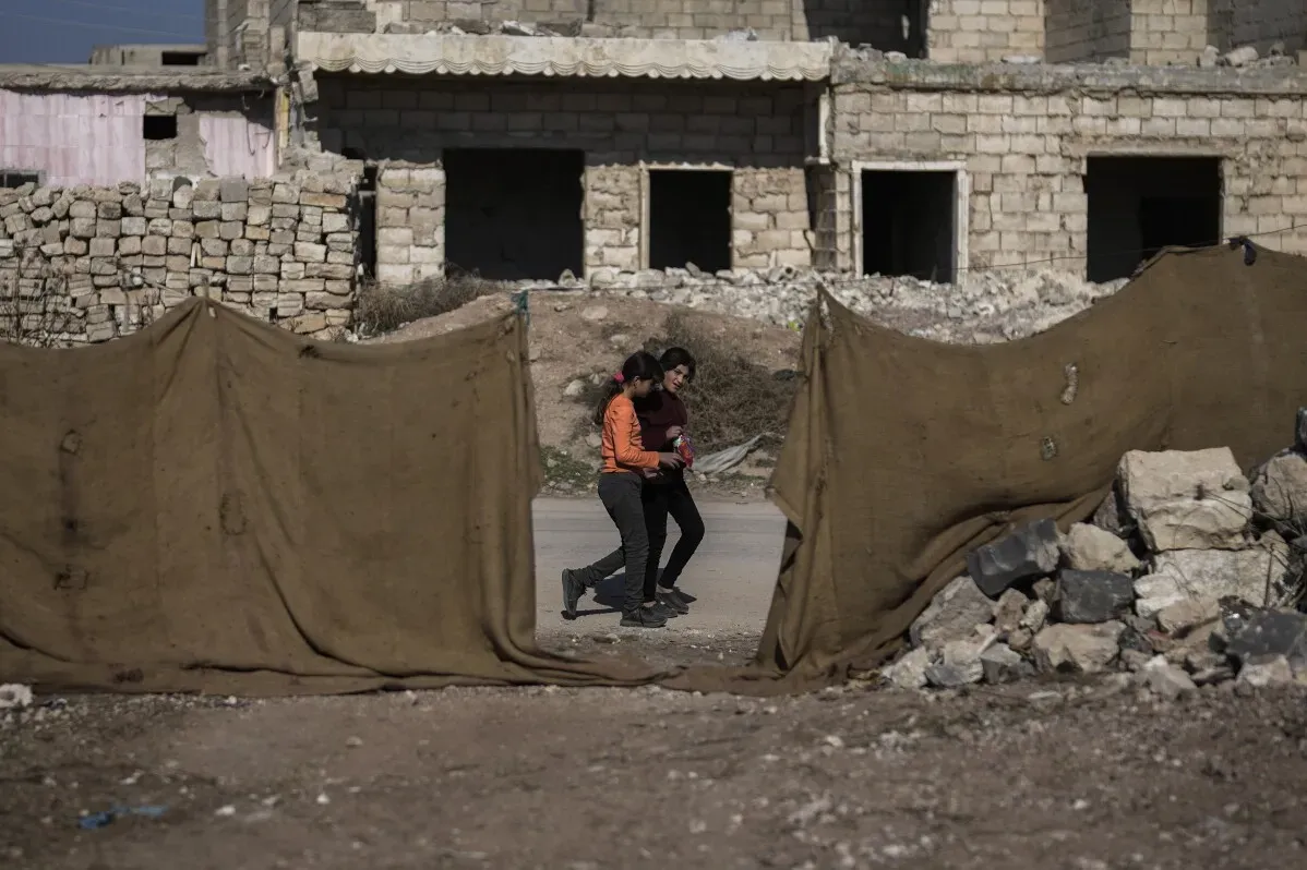 طفلتان تتجولان في شوارع تل رفعت المدمرة، محاطتين بالركام والأبنية غير المكتملة، تعكسان آثار النزاع المستمر في سوريا.