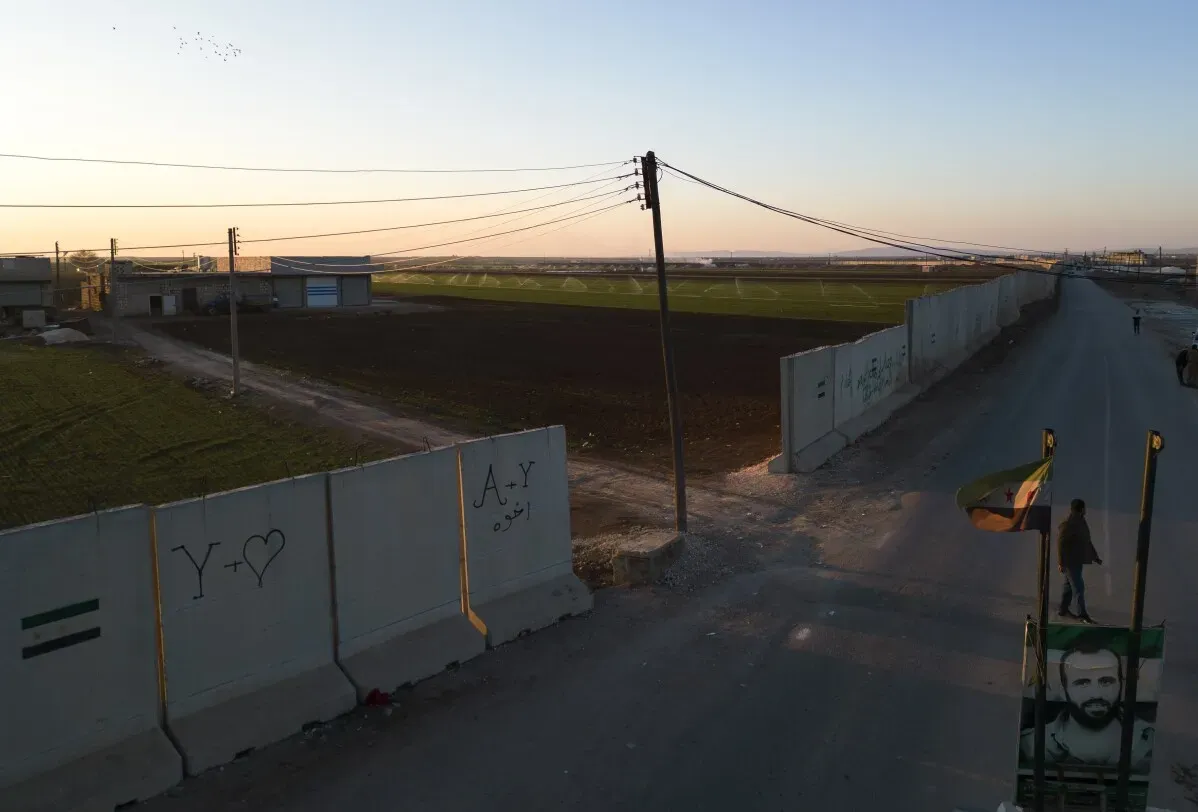 جدار خرساني يفصل بين تل رفعت والأراضي الزراعية، مع وجود آثار الحرب في الخلفية، تعكس تحديات إعادة الإعمار بعد النزاع.