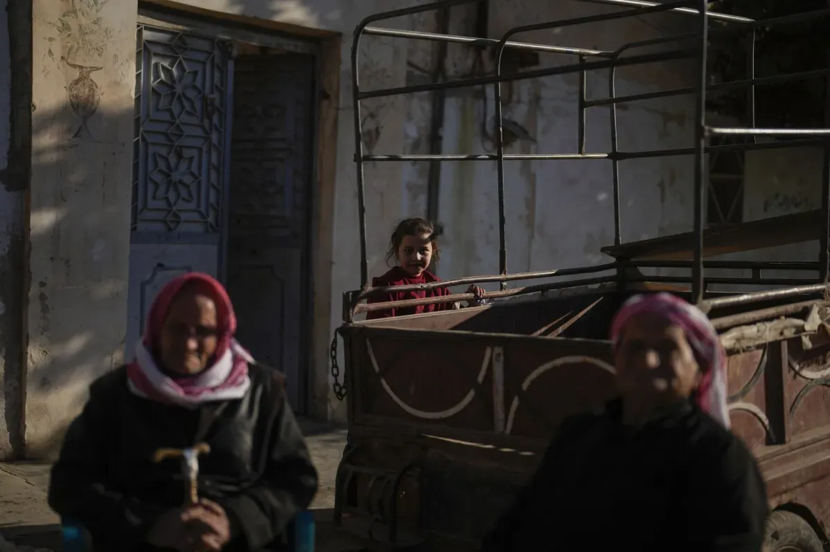 طفلة صغيرة ترتدي ملابس حمراء تقف خلف عربة قديمة، بينما تجلس امرأتان مسنّتان في المقدمة، في مشهد يعكس آثار الحرب في تل رفعت.