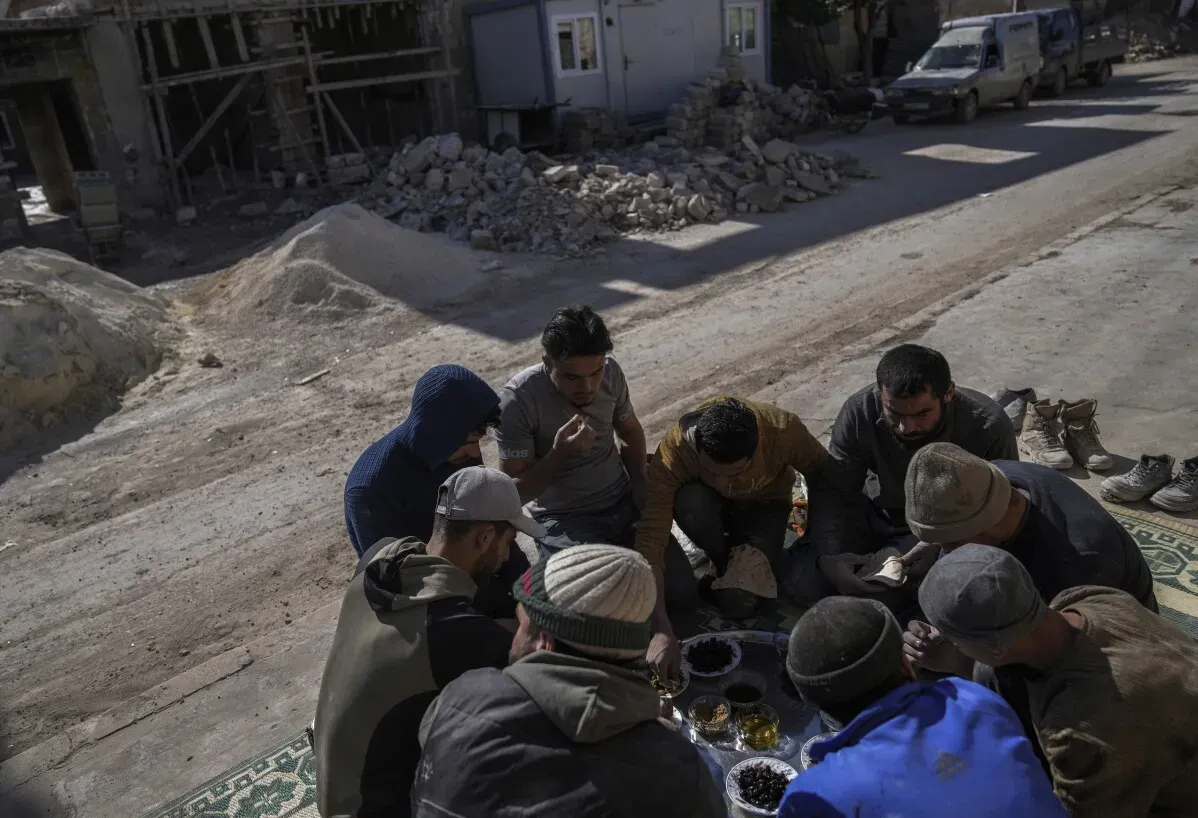 مجموعة من الرجال يجلسون على الأرض في تل رفعت، سوريا، يتناولون الطعام وسط أنقاض المباني المدمرة، مما يعكس تحديات إعادة الإعمار بعد الحرب.