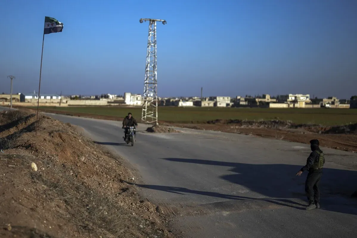 مشهد من تل رفعت يظهر رجلًا على دراجة نارية بالقرب من علم سوري، مع خلفية منازل متضررة وأراض زراعية، مما يعكس آثار النزاع المستمر.