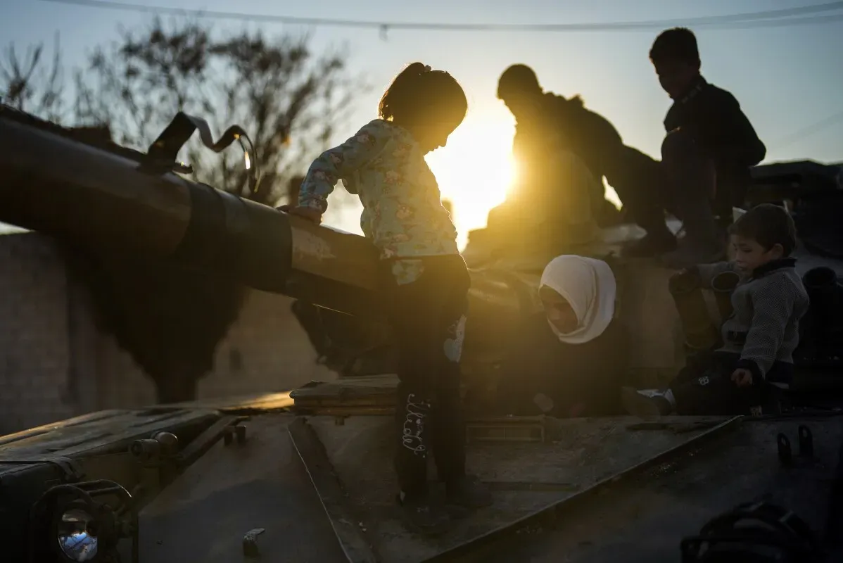 أطفال يلعبون على دبابة قديمة في تل رفعت، سوريا، مع غروب الشمس في الخلفية، مما يعكس آثار الحرب والعودة إلى الحياة اليومية.