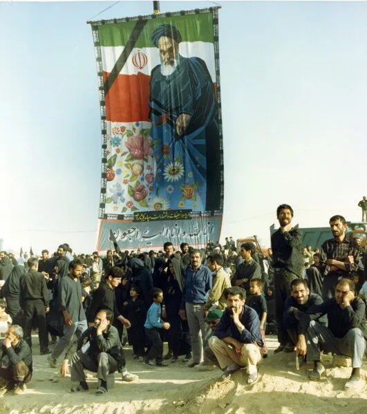 حشد كبير من المشيعين في جنازة آية الله روح الله الخميني عام 1989 أمام لافتة ضخمة تحمل صورته وعلم إيران، في مشهد تاريخي من الحزن والتجمع الشعبي.
