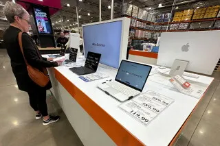 امرأة تتفقد أجهزة Mac وiPad في متجر، مع لافتات توضح الأسعار الجديدة بعد الزيادة، في سياق أزمة نقص رقائق الذاكرة.