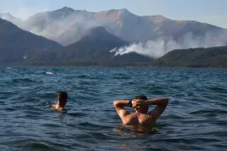 دخان حرائق الغابات يتصاعد من جبال باتاغونيا في الأرجنتين، بينما يسبح رجلان في بحيرة، مع مشهد مروع في الخلفية.
