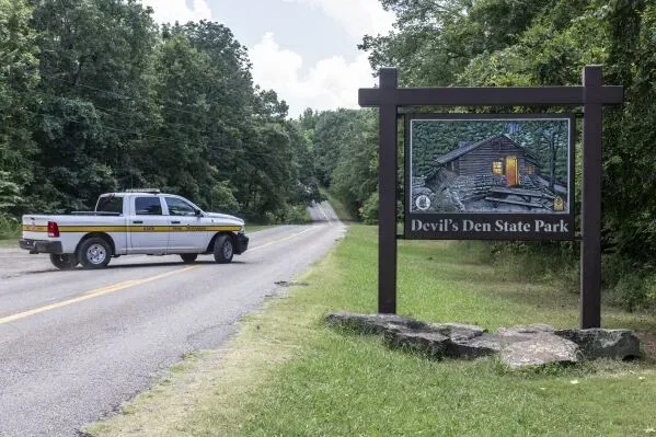 لافتة تشير إلى متنزه Devil's Den State Park، مع شاحنة شرطة متوقفة على جانب الطريق وسط طبيعة خضراء.