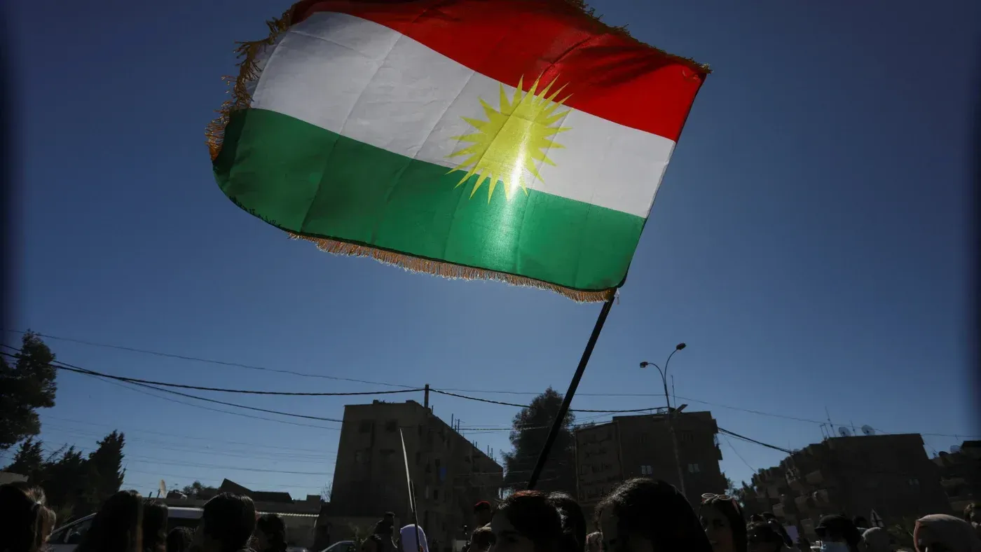 علم كردي يرفرف في الهواء، مع خلفية لمباني في منطقة حضرية، يعكس التوترات السياسية والجهود الكردية ضد النظام الإيراني.