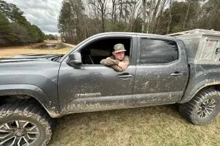 رجل يجلس في نافذة سيارة بيك أب مظللة، محاطًا بأشجار، مما يعكس أجواء الريف. السيارة مغطاة بالطين، مما يشير إلى نشاط خارجي.